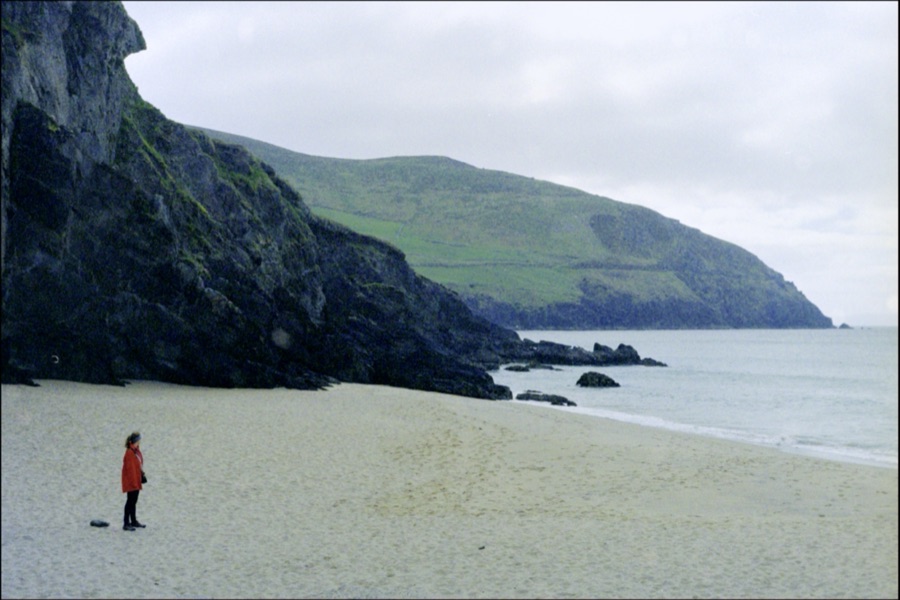 A person in a red coat alone on an empty sand beach below green headlands