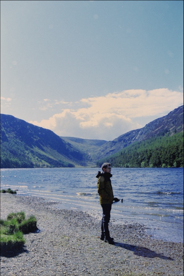 A person standing on the stony shore of a lake between steep green valley sides