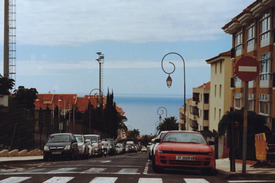 A red car parked on a street sloping down towards the sea